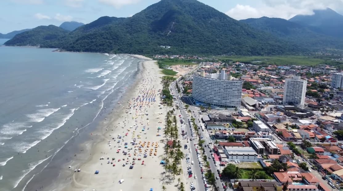 Vista aérea do litoral de Peruíbe, com o morro coberto de mata à esquerda, a faixa de praia, a avenida arborizada à beira-mar e a cidade à direita.